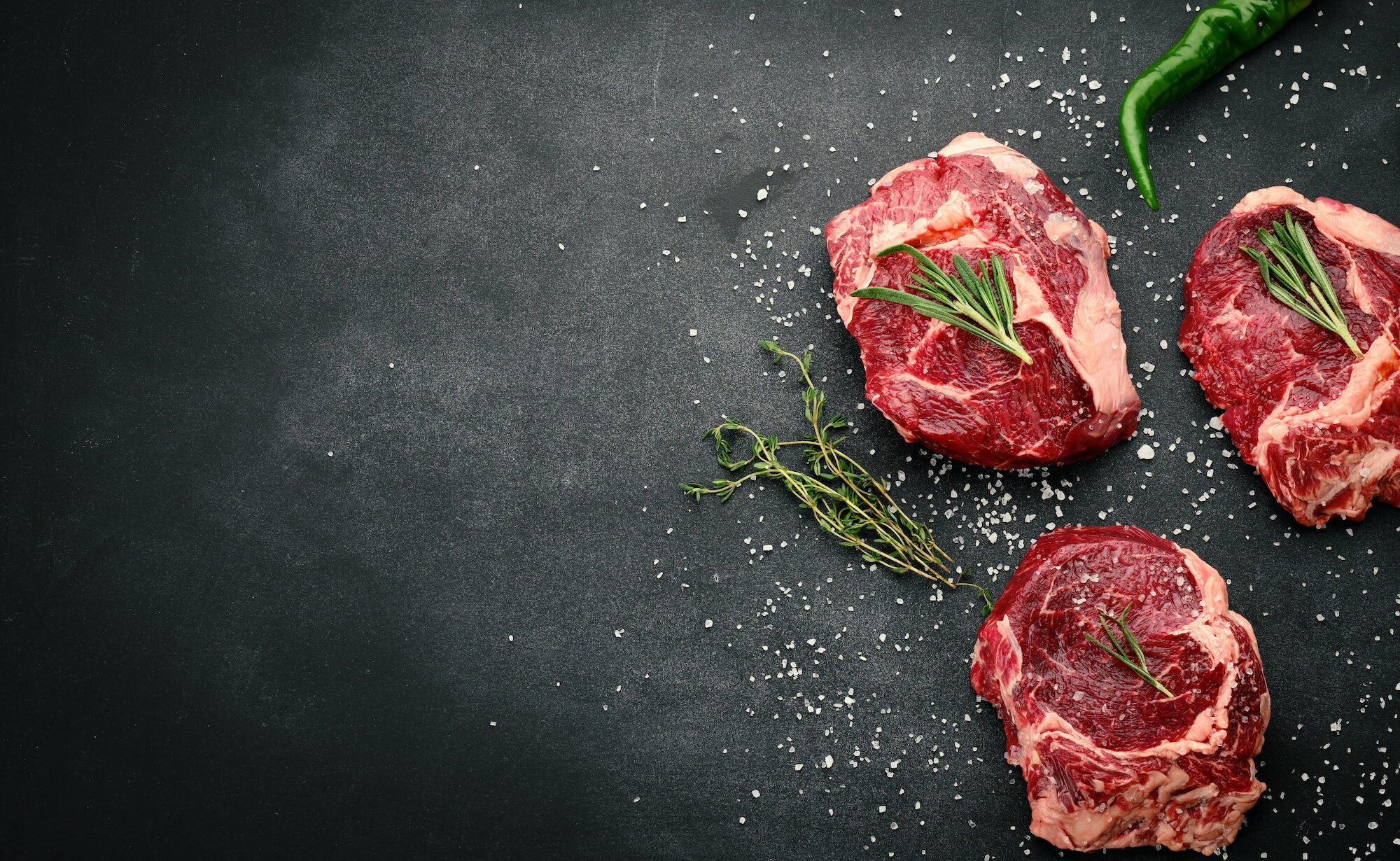 Raw piece of beef ribeye with rosemary, thyme on a black table, top view